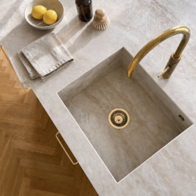 Dettaglio dall’alto di un lavello in pietra chiara con rubinetto in ottone, parquet a spina e accessori da cucina in materiali naturali.